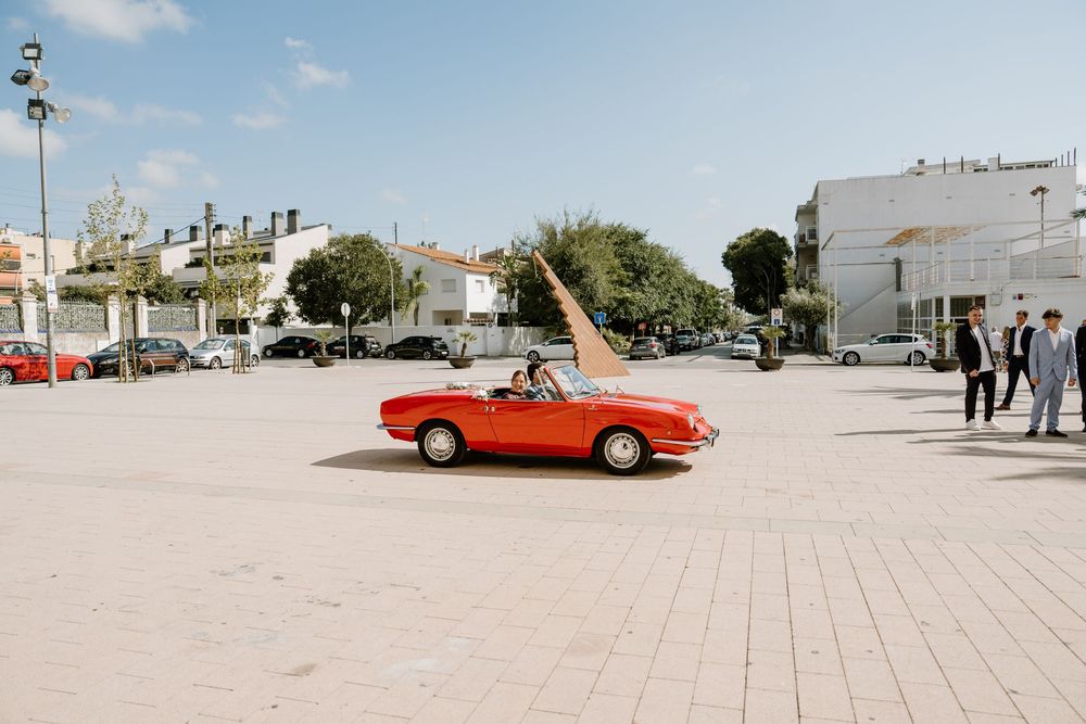 Red vintage convertible car parked on a sunny paved plaza with people standing nearby and buildings and trees in the background.