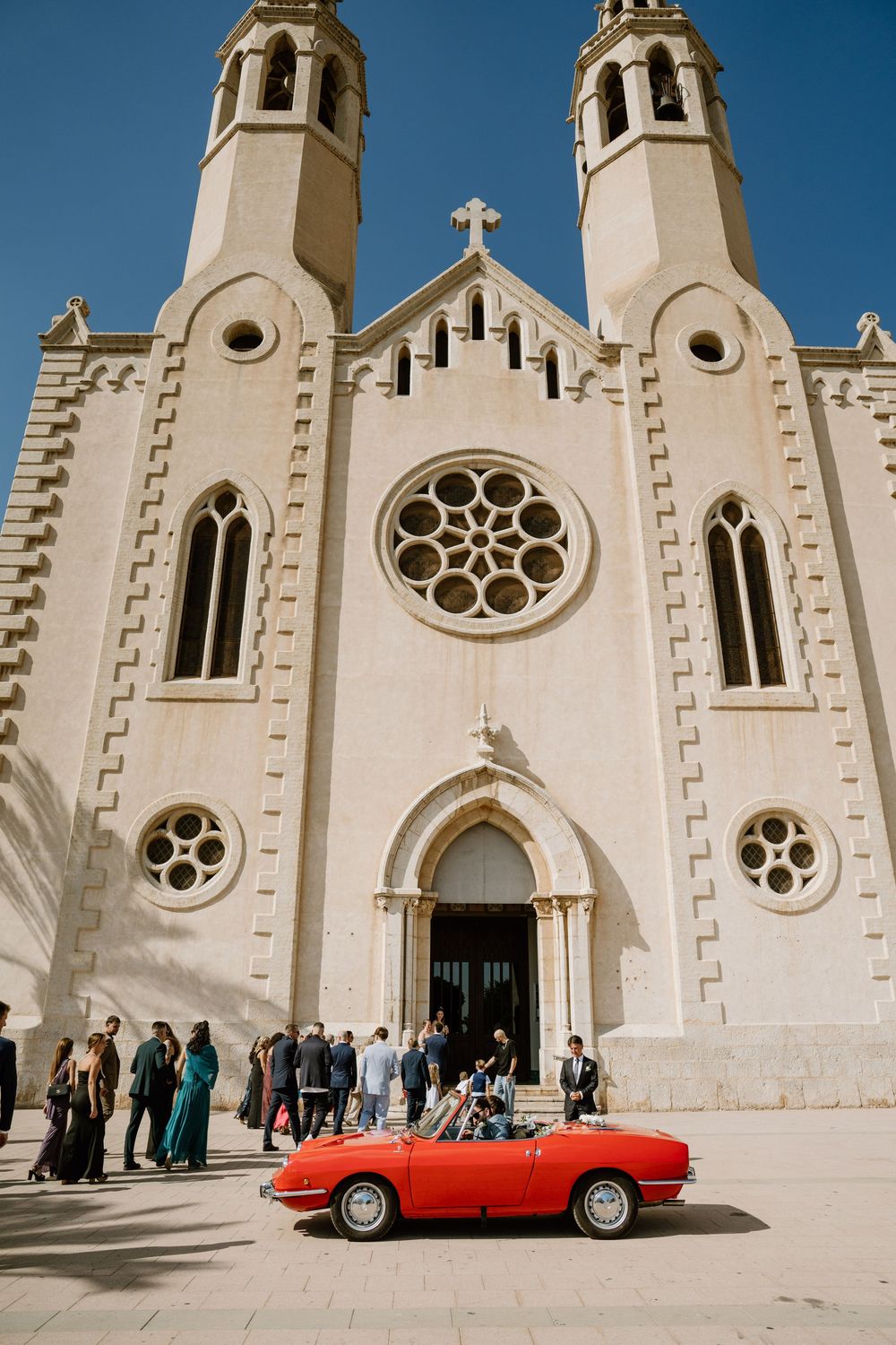 Wedding guests gather outside a cream stone church with twin bell towers and a rose window, beside a red vintage convertible.