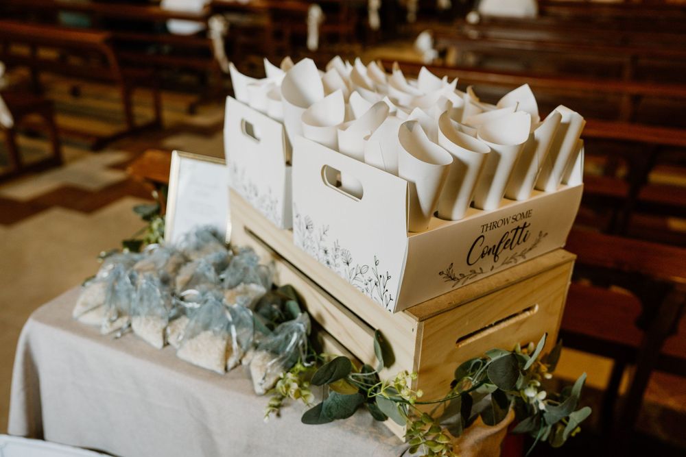 White paper cones filled with confetti in a labeled wooden box on a table with eucalyptus greenery, ready for a church wedding ceremony.