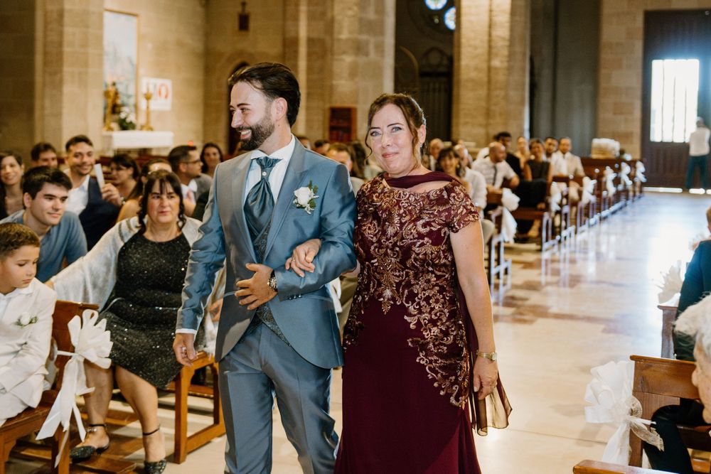 Smiling man in a blue suit and woman in a burgundy lace dress walk arm in arm down a church aisle past seated wedding guests.