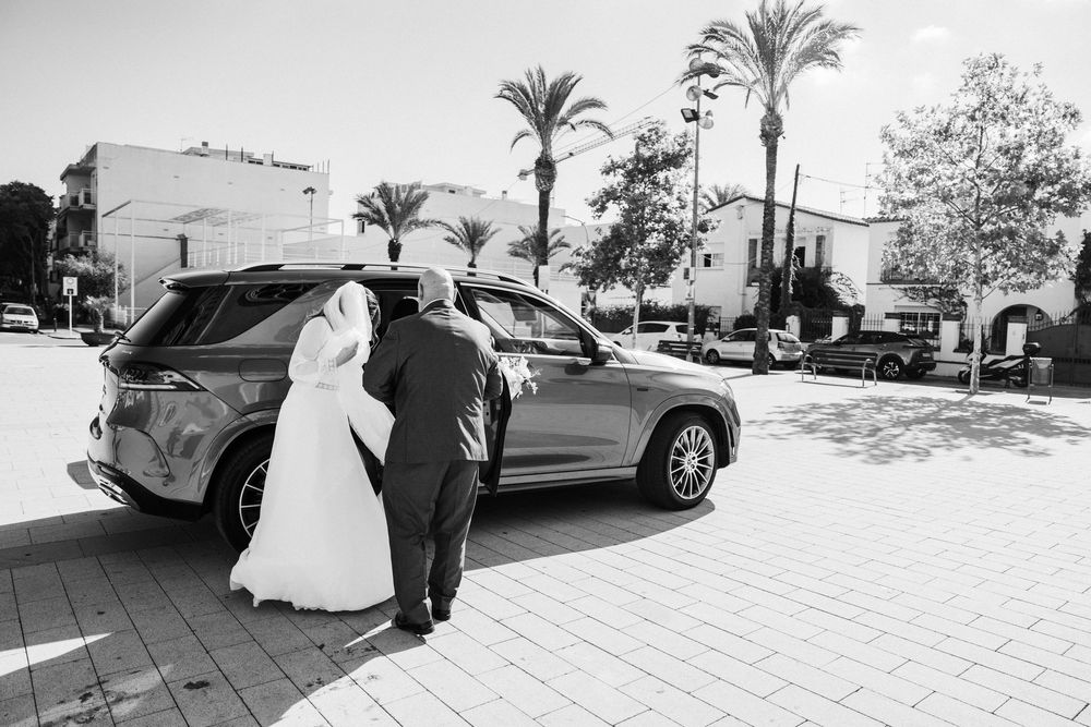 Bride in a white wedding gown and veil walks with a man in a dark suit toward an SUV on a sunlit palm-lined plaza in black and white.