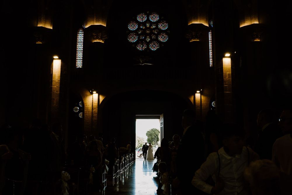 Silhouetted wedding guests fill a dimly lit church as the bride and groom enter through the bright doorway beneath a rose window.