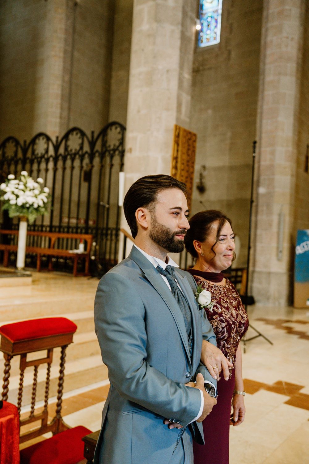 Groom in a light blue suit with a floral boutonniere stands beside a smiling woman in a burgundy dress inside a stone church.