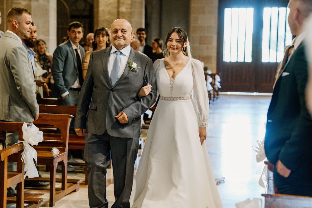 A bride in a white wedding gown walks down the church aisle arm in arm with an older man in a gray suit as guests look on.