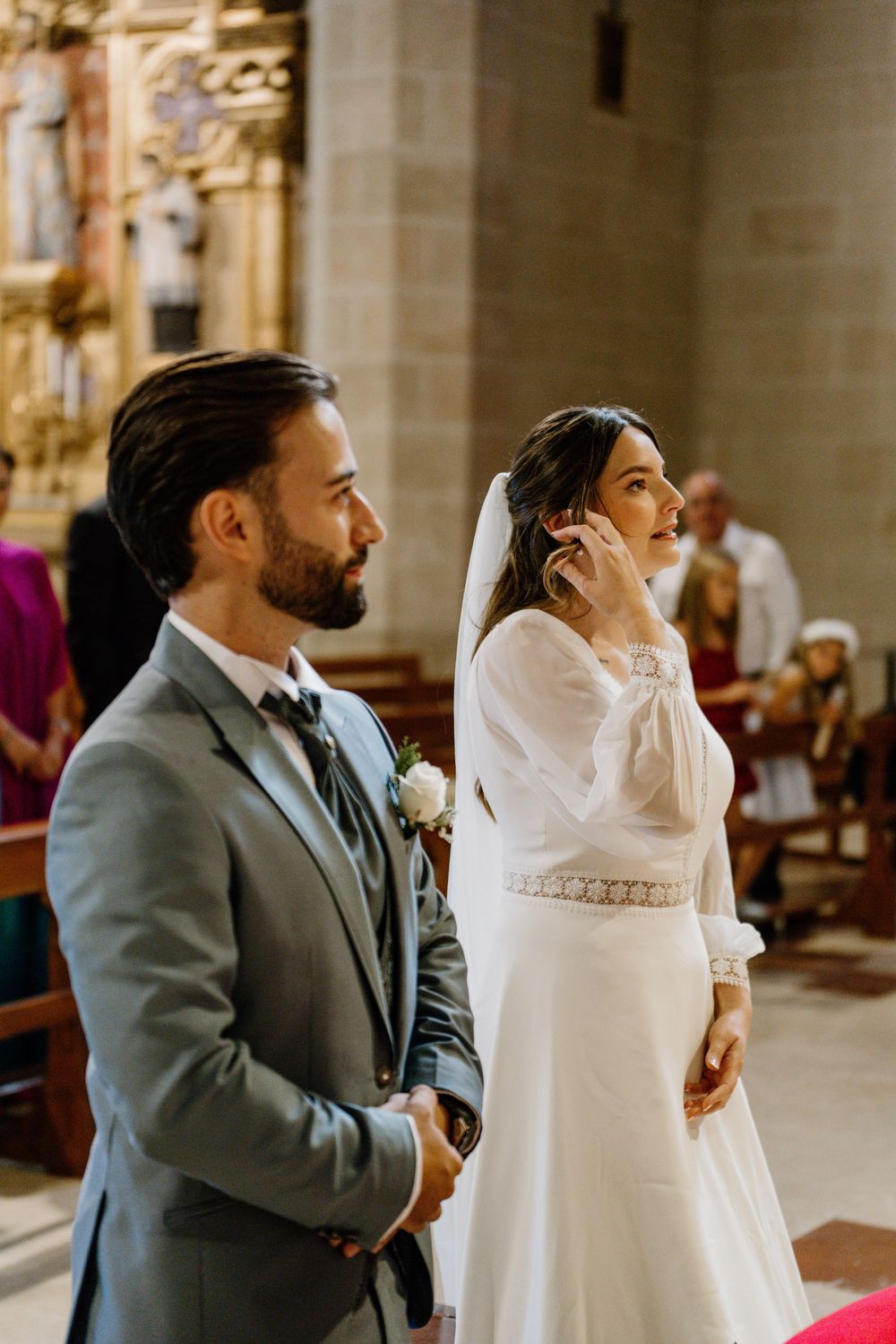 Bride in a white long-sleeve gown and groom in a gray suit with boutonniere stand together during a church wedding ceremony.
