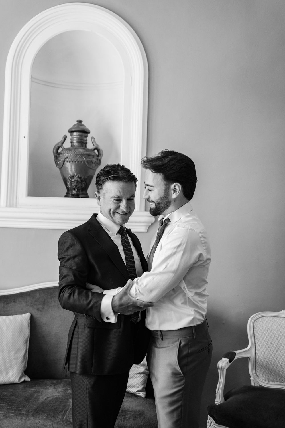 Two men in dress shirts and ties share a warm, smiling embrace in a bright room with an arched wall niche and vase.
