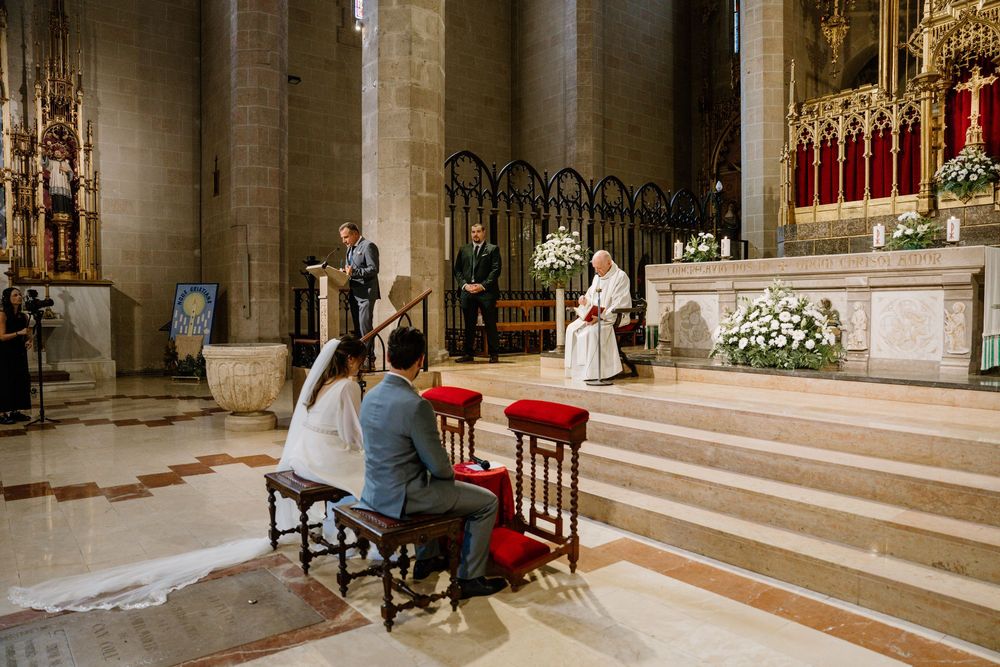Bride and groom kneel on red stools before a priest at the altar during a wedding ceremony inside a historic stone church.
