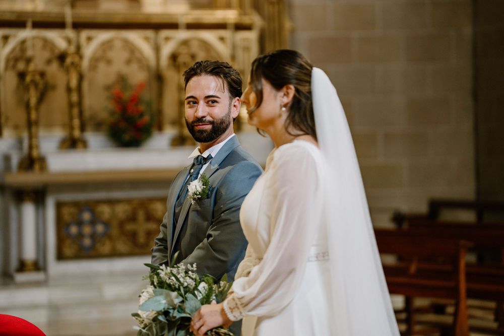 Smiling groom in a gray suit with a boutonniere gazes at his bride in a white gown and long veil during a church wedding ceremony.