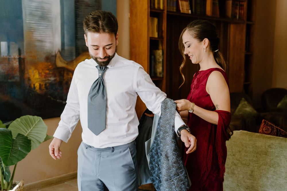 A woman in a red dress helps a smiling bearded man in a white shirt and gray tie put on a patterned jacket in a bright, cozy room.