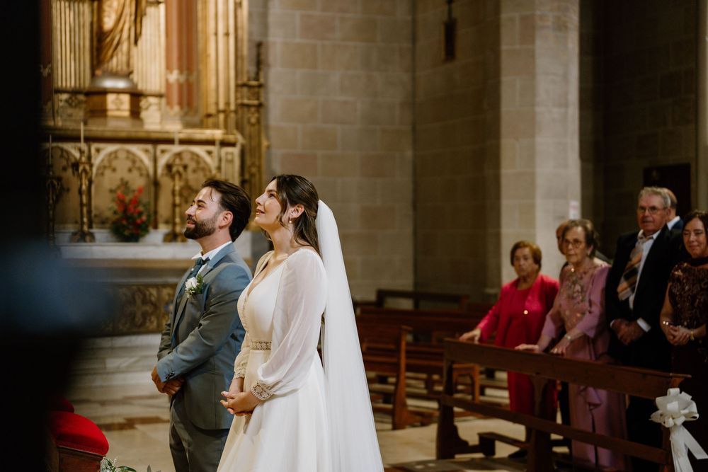 Bride in a white gown and veil stands beside the groom in a gray suit during a church wedding ceremony as guests watch from the pews.
