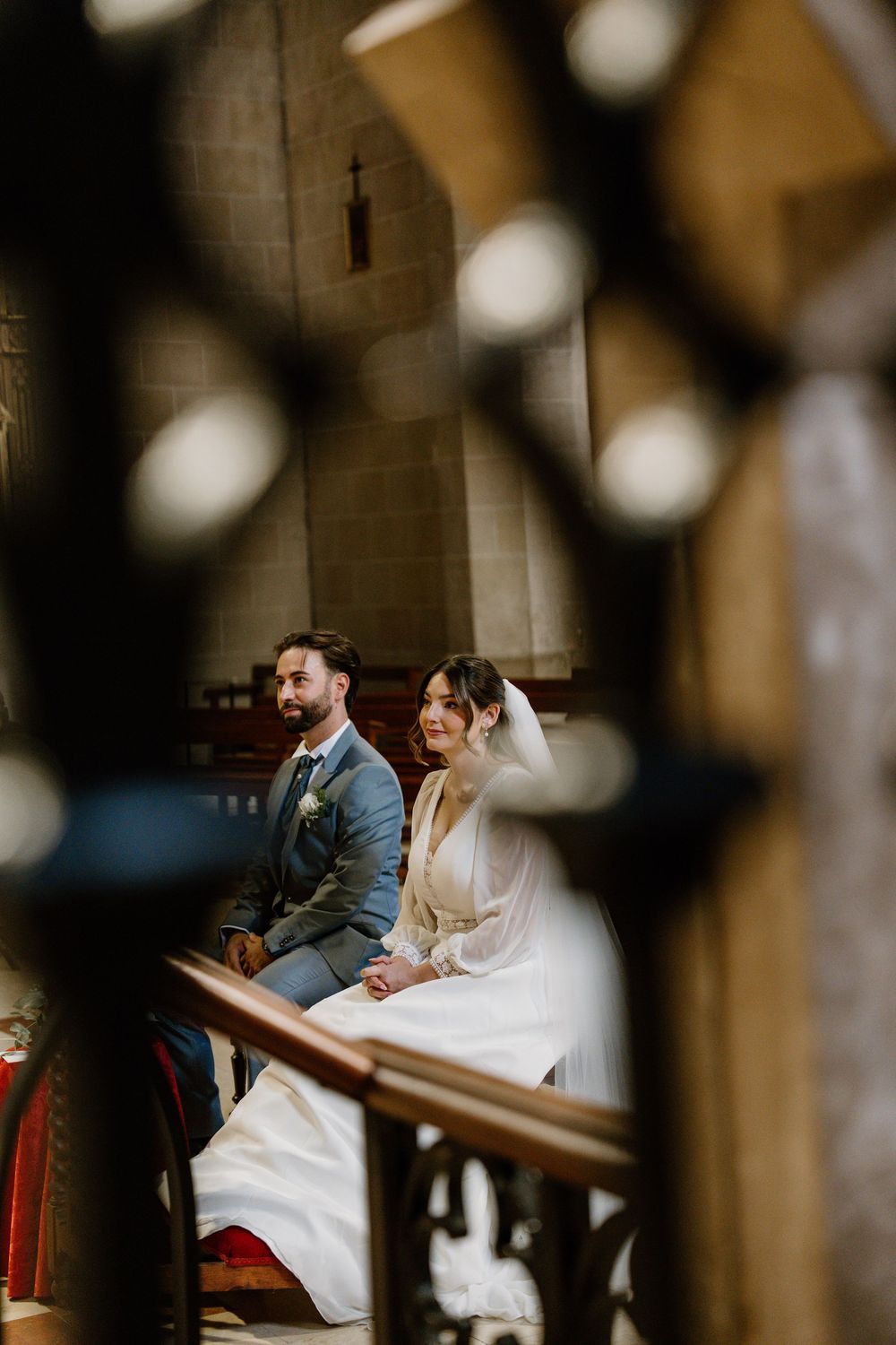 Bride in a white gown and groom in a gray suit sit together in a stone church during their wedding ceremony, framed by ornate ironwork.