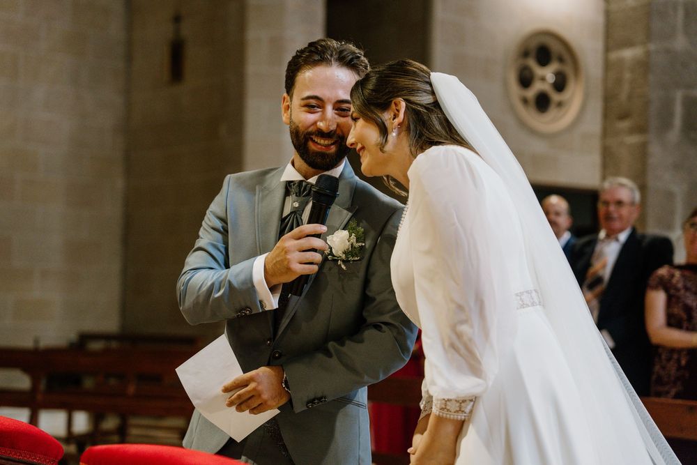 Smiling groom in a gray suit speaks into a microphone while the bride in a white veil laughs beside him during a church wedding ceremony.