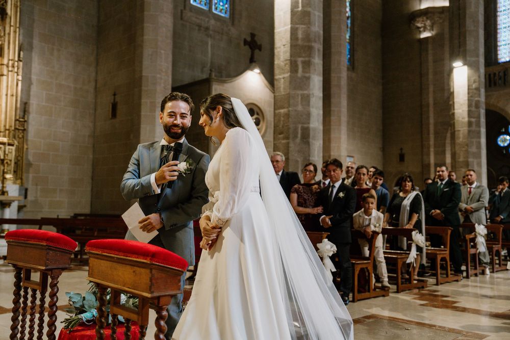 Groom in a gray suit smiles while holding a microphone beside a bride in a long veil during a wedding ceremony in a stone church.