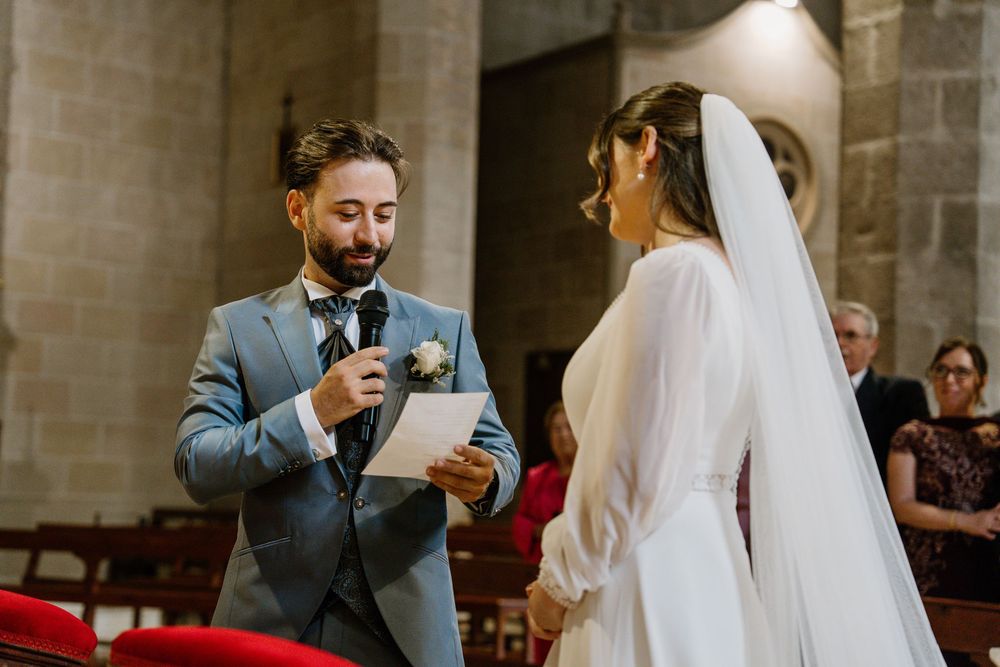 Groom in a gray suit reads his handwritten vows into a microphone as the bride in a long veil listens at a church wedding.