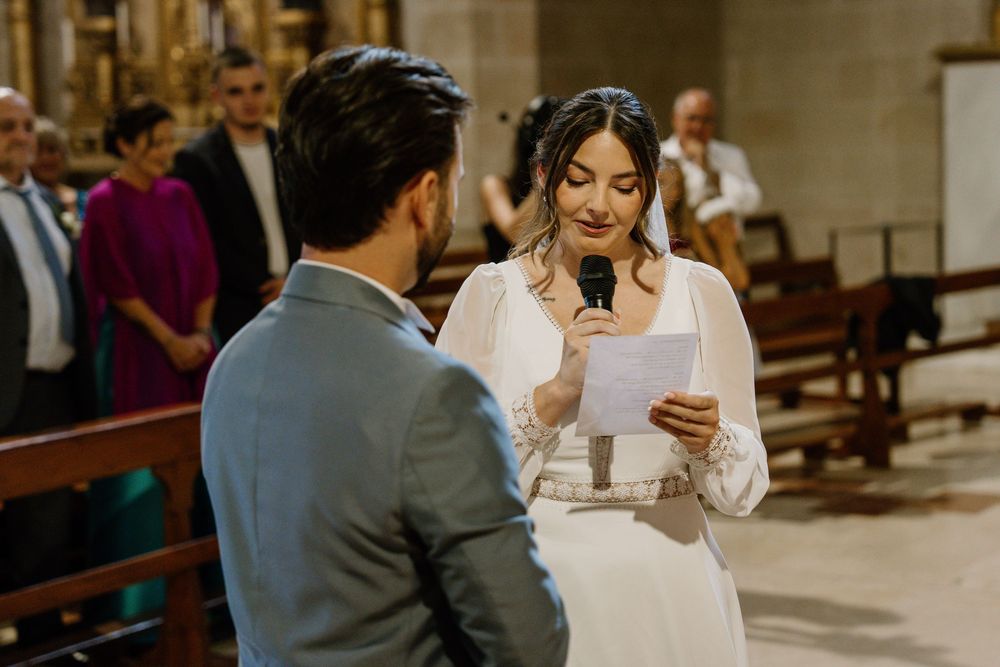 Bride in a white dress reads from a paper into a microphone during a church wedding ceremony as the groom and guests look on.