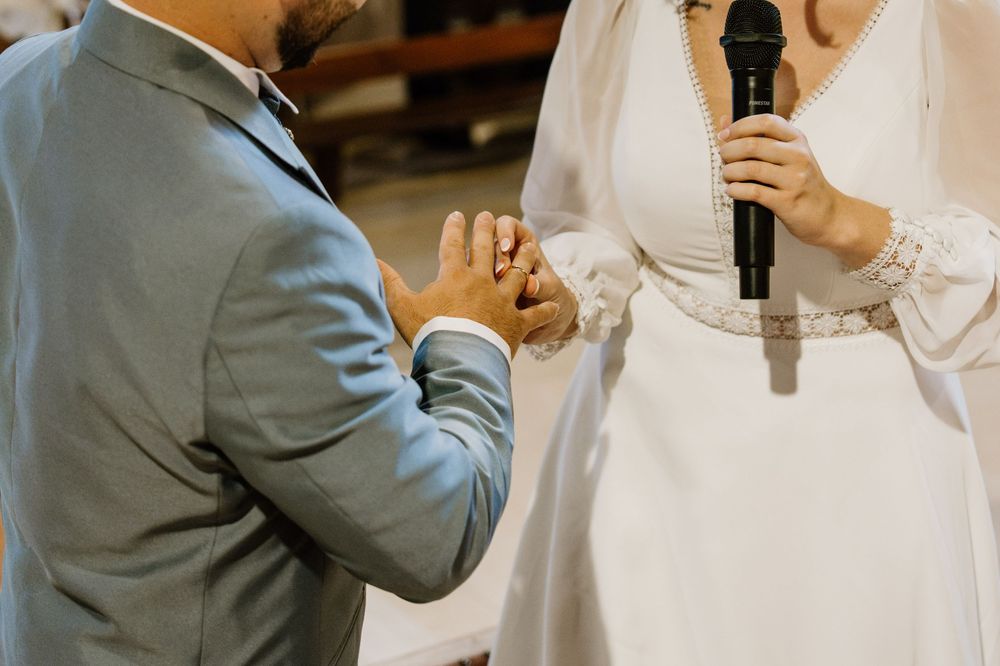 Groom in a gray suit places a ring on the bride's finger as she holds a microphone during a wedding ceremony.