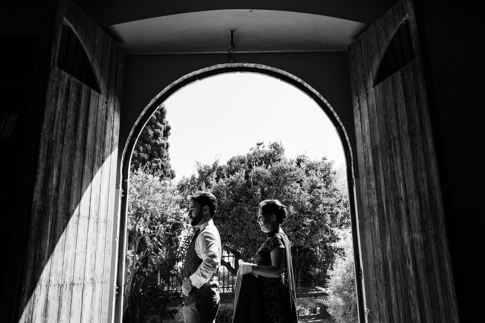 Black and white photo of a couple silhouetted in a large arched doorway, framed by wooden doors and sunlit trees in the background.
