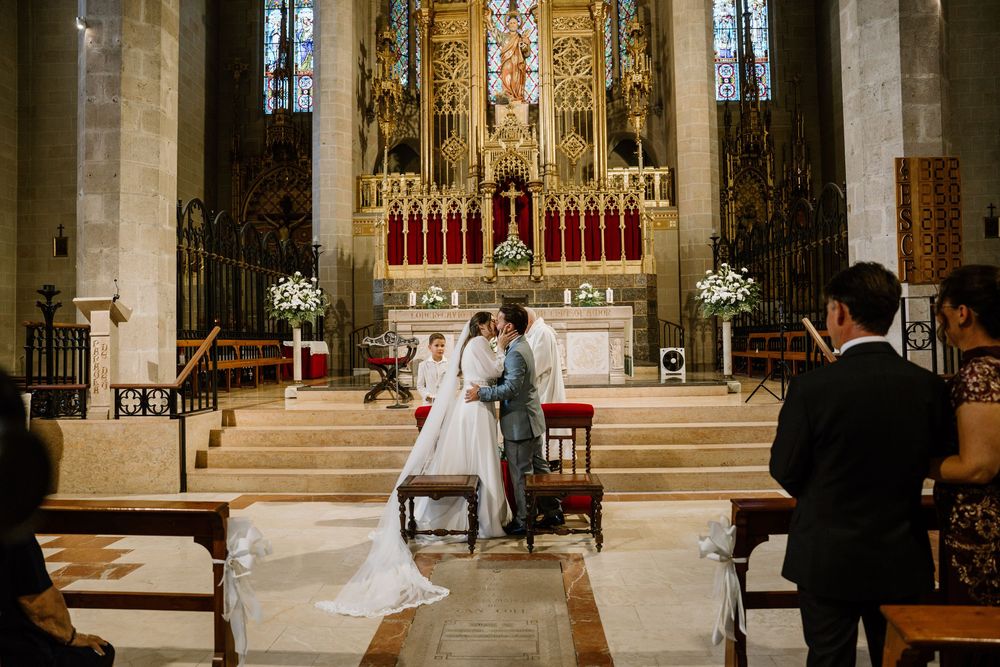 Bride in a white gown and groom kneel at the altar during a wedding ceremony inside an ornate church with stained glass windows.