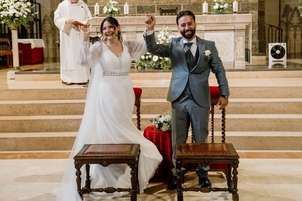 Joyful bride in a white lace gown and groom in a gray suit raise their fists in celebration after their church wedding ceremony.