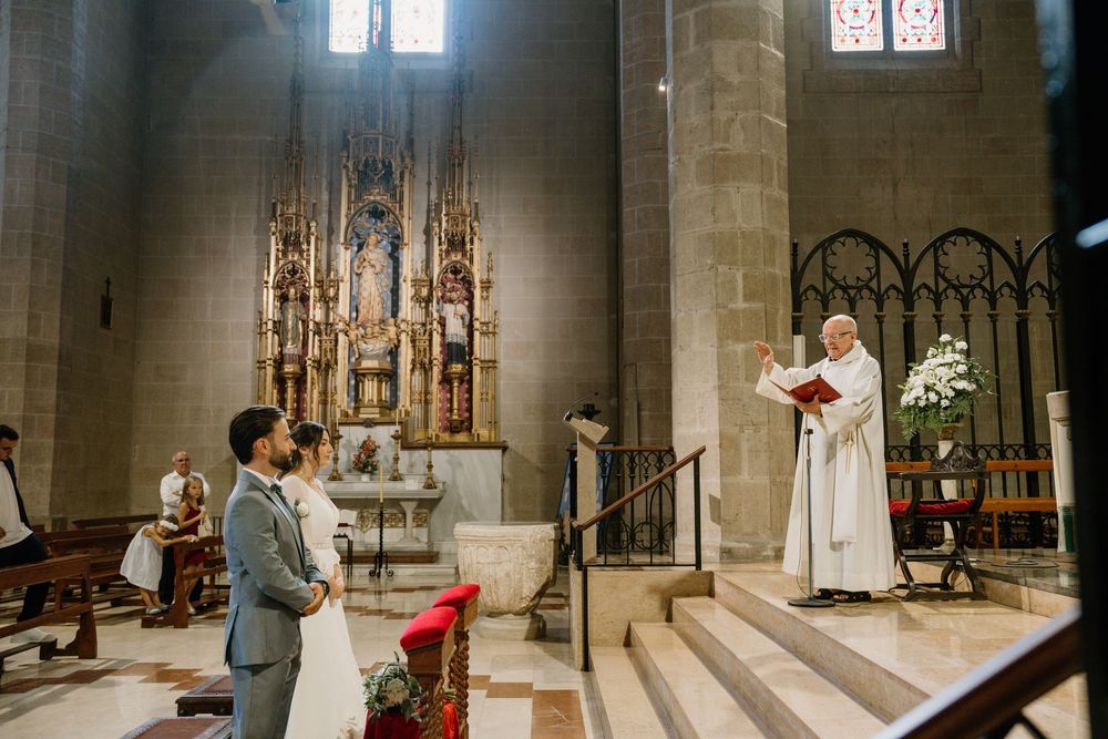 Bride and groom stand before a priest holding a red book during a wedding ceremony inside a stone church with an ornate golden altar.