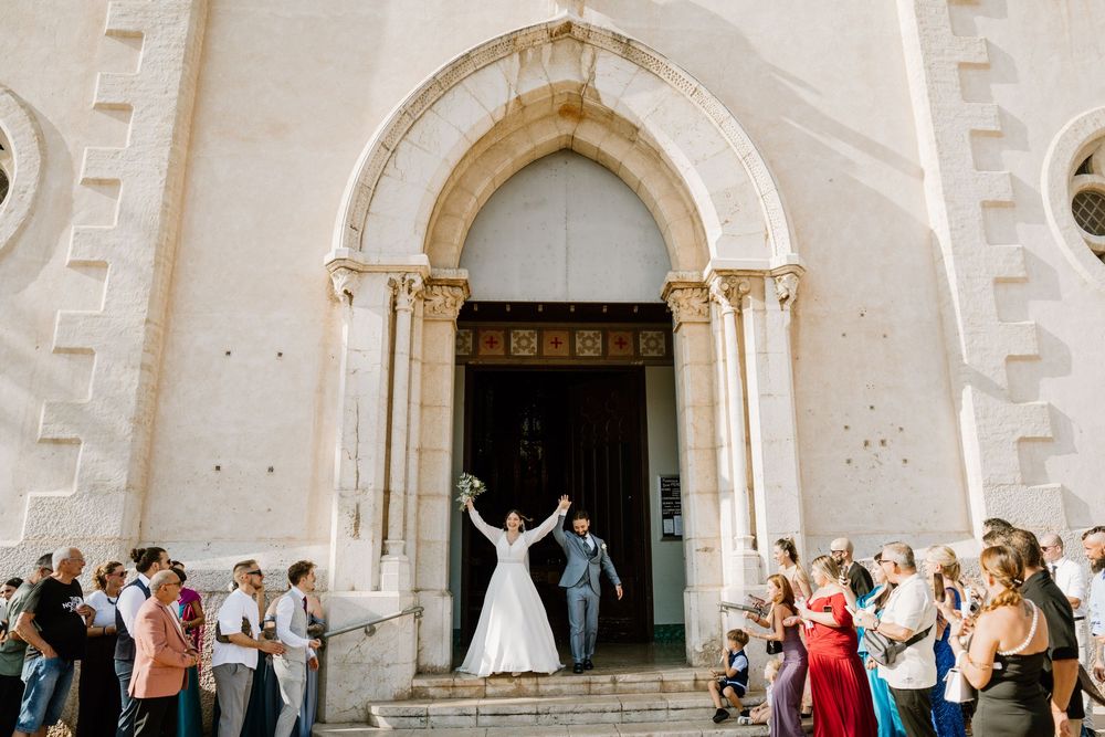 Bride and groom raise their arms in celebration as they exit a stone church while wedding guests cheer from the steps below.