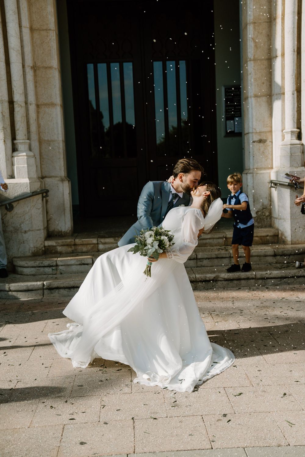 Groom in a blue suit dips and kisses his bride in a white gown on church steps as a boy watches and confetti falls.