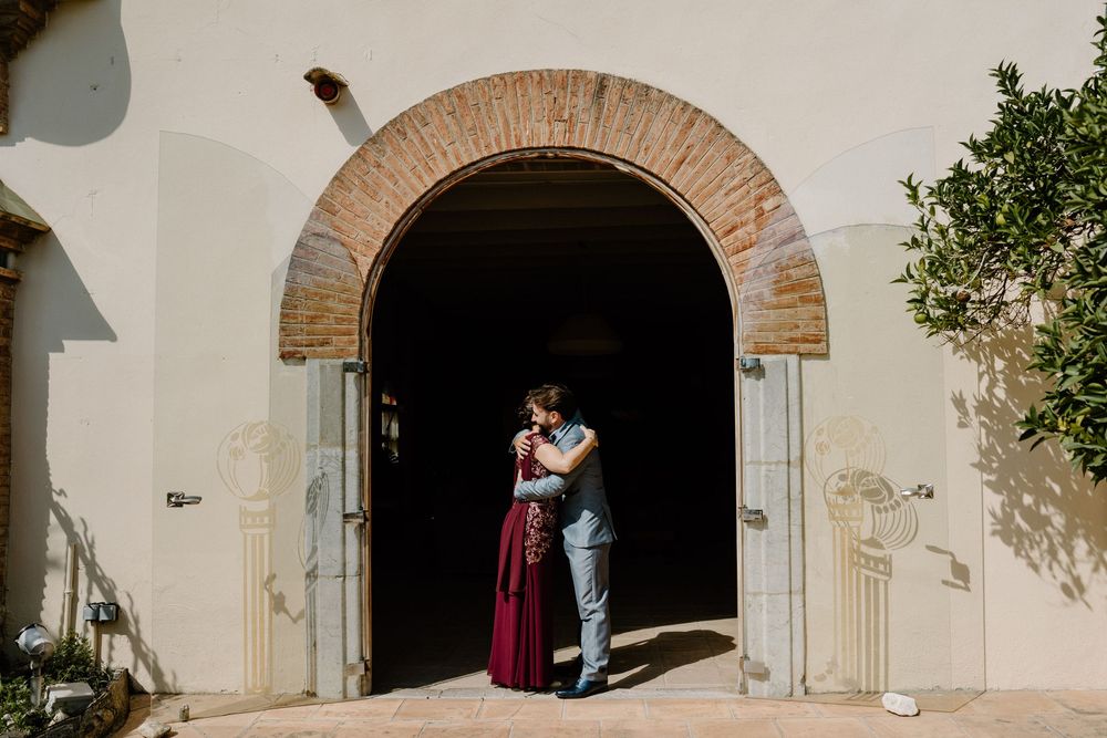 Couple embracing in front of a rustic brick archway entrance, she wears a long burgundy skirt, on a sunlit stone patio beside green foliage.