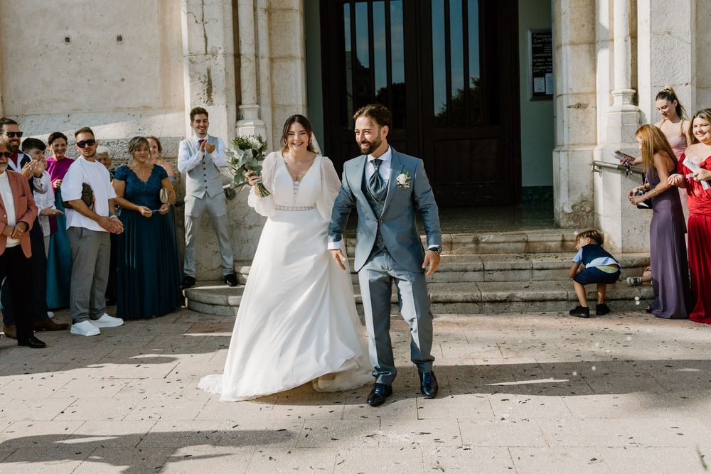 Bride in a white gown and groom in a gray suit walk hand in hand outside a stone church while wedding guests look on.