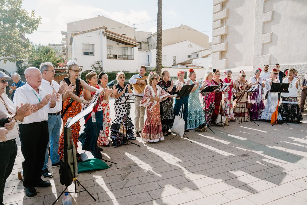 Group of people in colorful traditional dresses sing and clap together on a sunny paved plaza with music stands and white buildings behind.