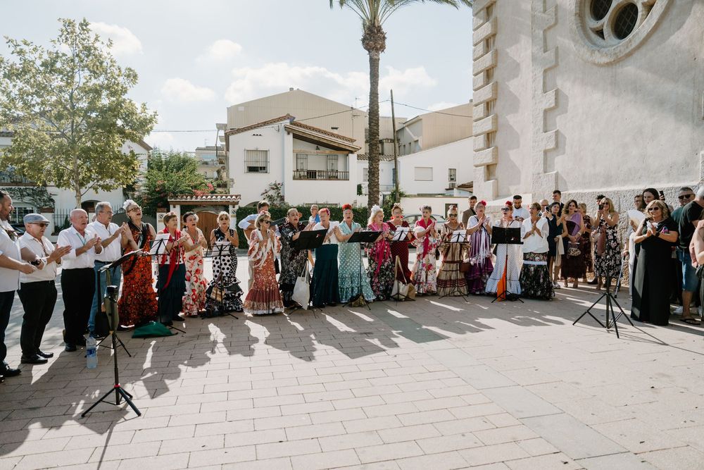 Choir in colorful traditional costumes sings outdoors on a sunny plaza with microphones, a palm tree and a stone church in the background.