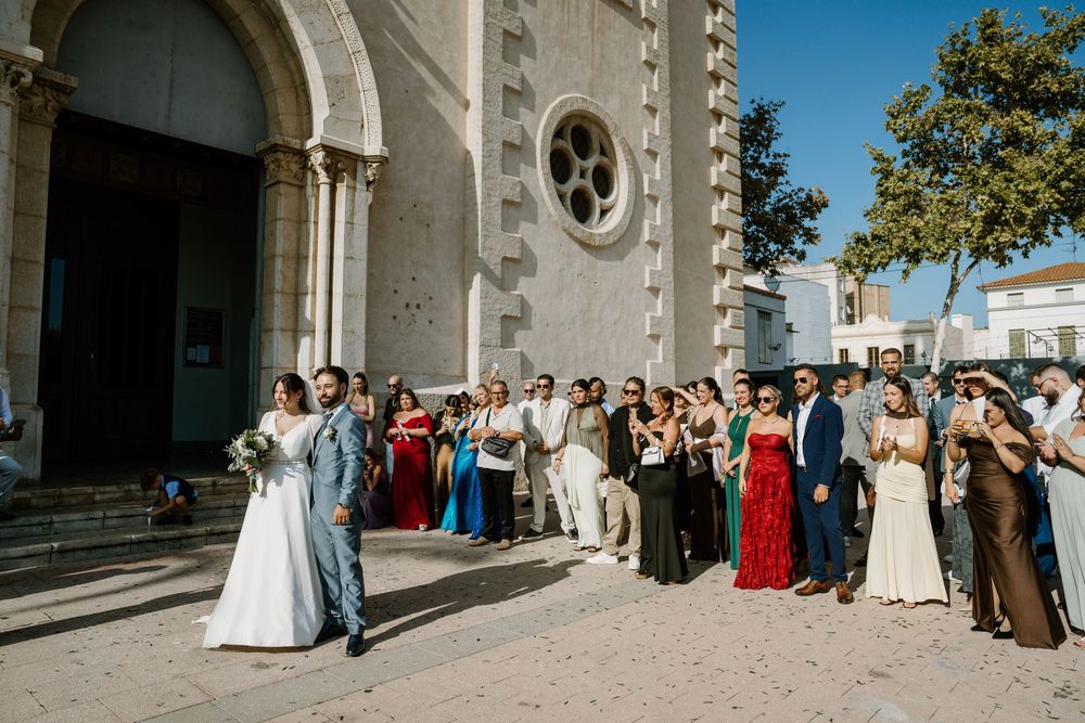 Bride in a white gown and groom in a blue suit walk past wedding guests lined up outside a historic stone church on a sunny day.