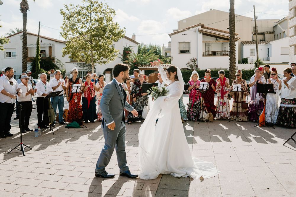 Bride in a white gown holding a bouquet and groom in a gray suit stand in a sunny plaza as guests in colorful traditional attire watch.