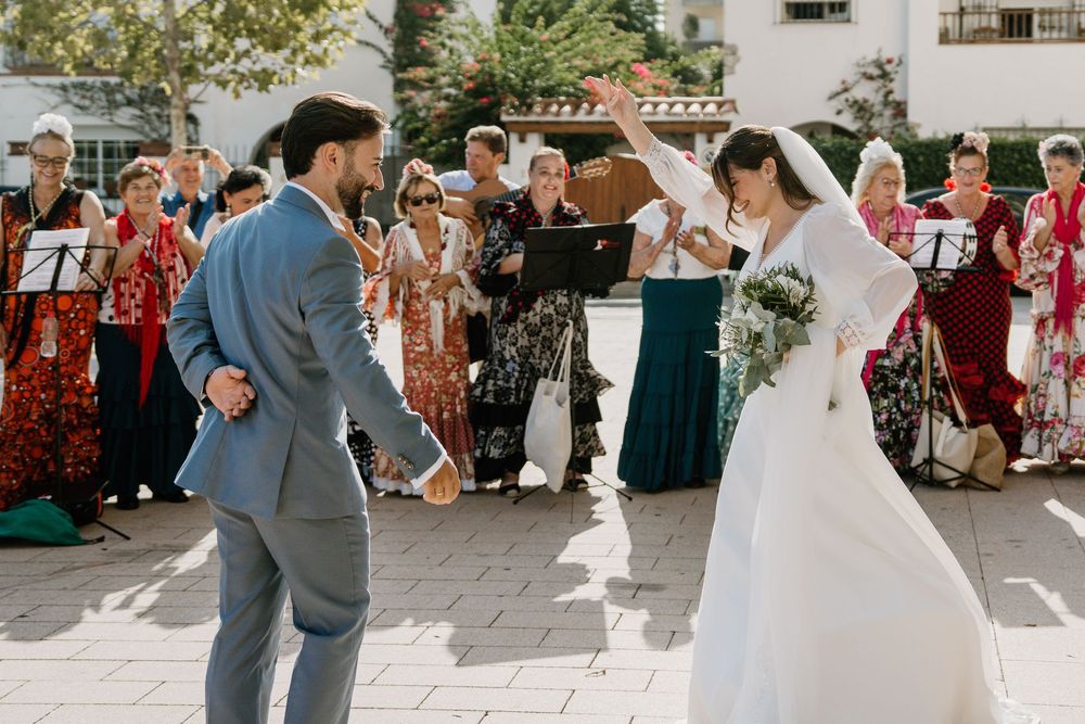 Bride in a white gown holding a bouquet dances with a groom in a gray suit as musicians and wedding guests watch at an outdoor ceremony.