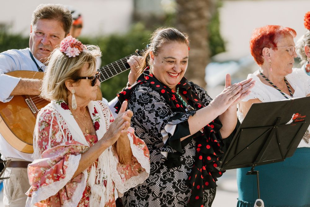 Smiling women in flamenco-style ruffled dresses clap along as a guitarist plays at a lively outdoor Spanish music performance.