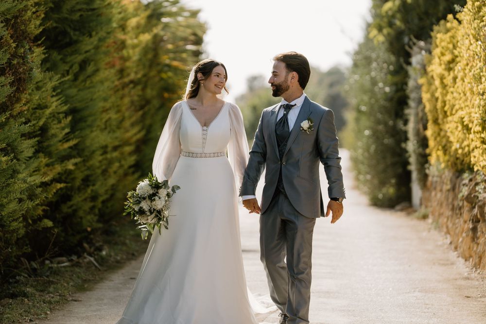 Bride in a flowing white gown holding a bouquet walks hand in hand with a groom in a gray suit along a sunlit path lined with green hedges.