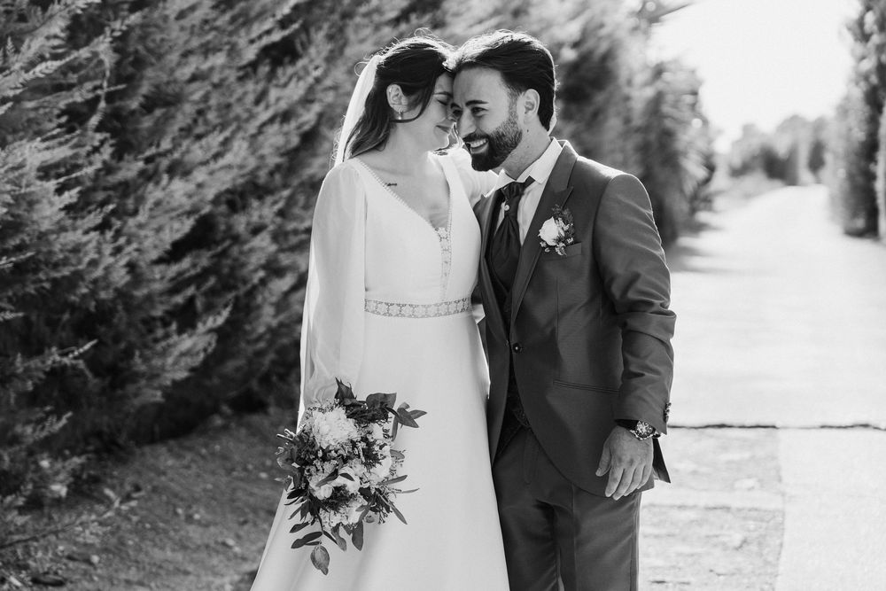 Black and white photo of a smiling bride and groom touching foreheads on a tree-lined path, the bride holding a floral bouquet.