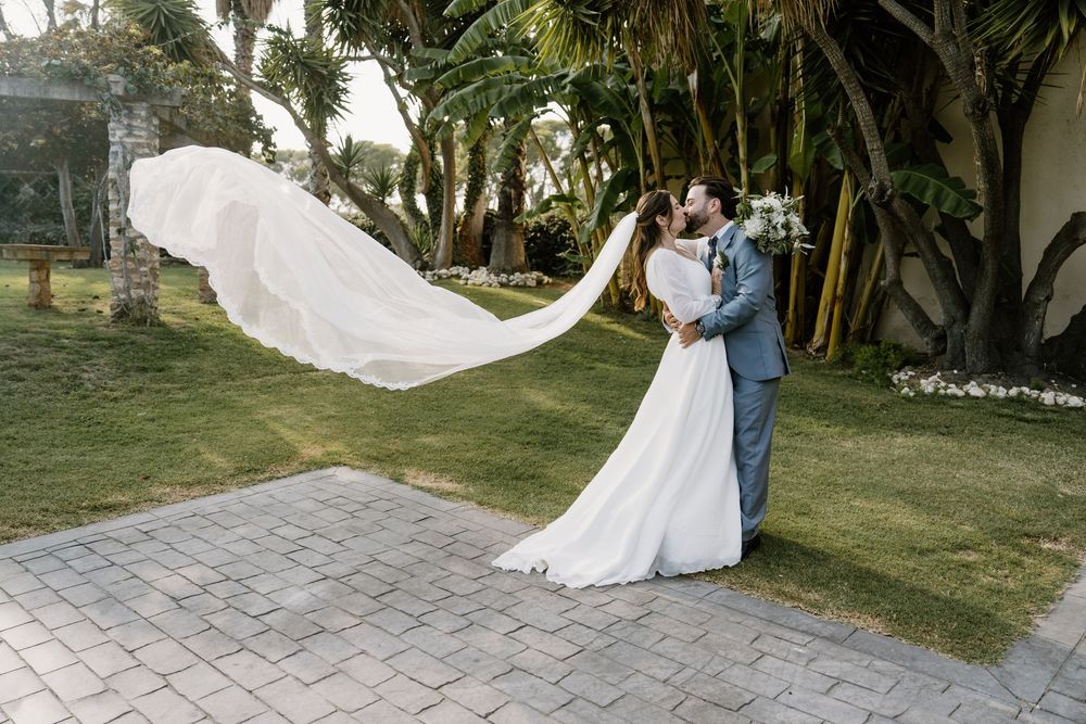 Bride in a flowing white gown and groom in a gray suit embrace on a lawn as her long veil billows in the wind among palm trees.