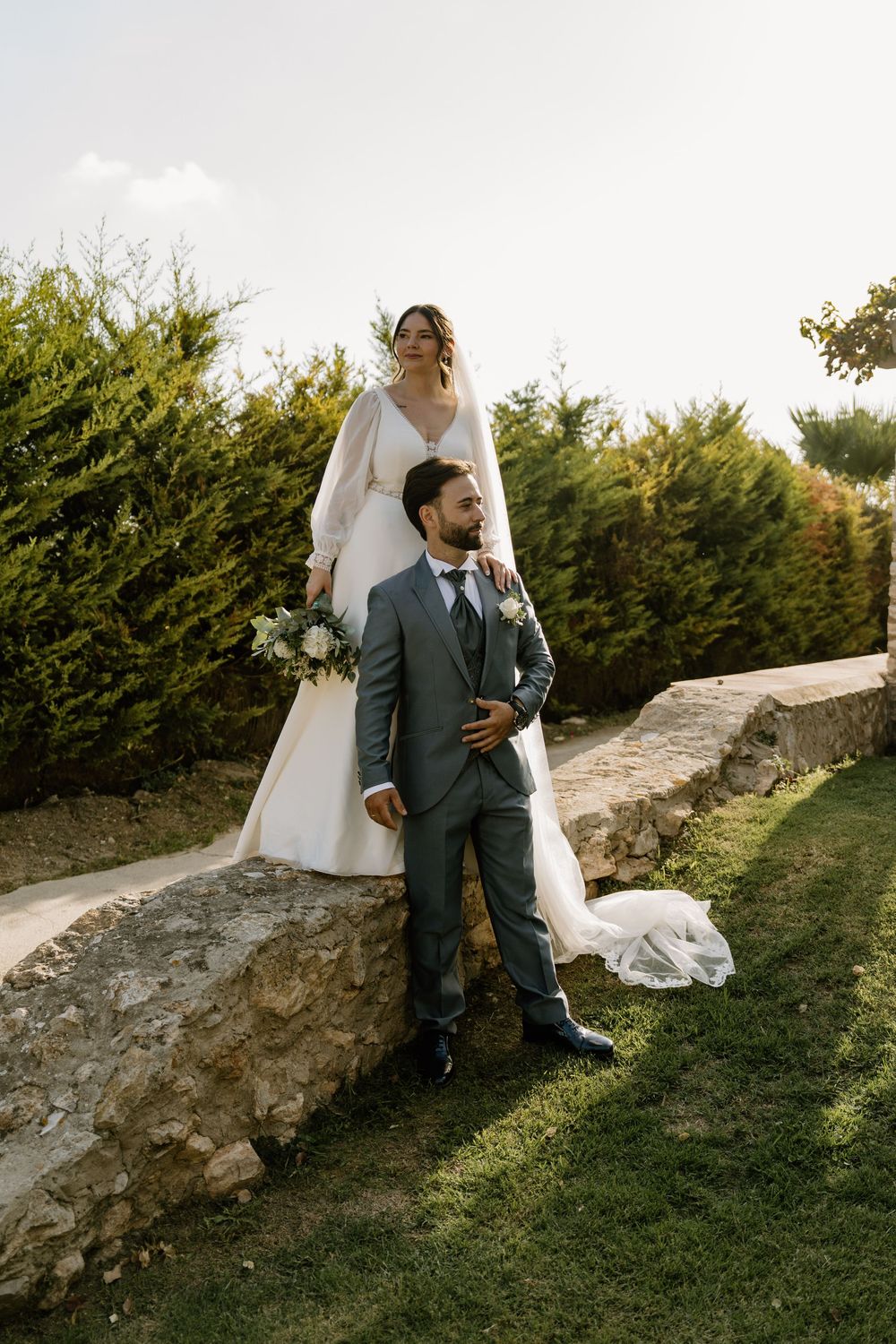 Bride in a flowing white gown holding a bouquet stands on a stone wall behind her groom in a gray suit on a sunny lawn.
