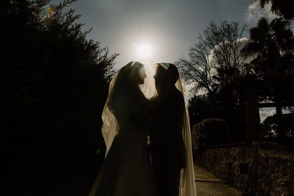 Silhouetted bride and groom stand close beneath a sheer veil as the sun glows behind them along a tree-lined stone path.