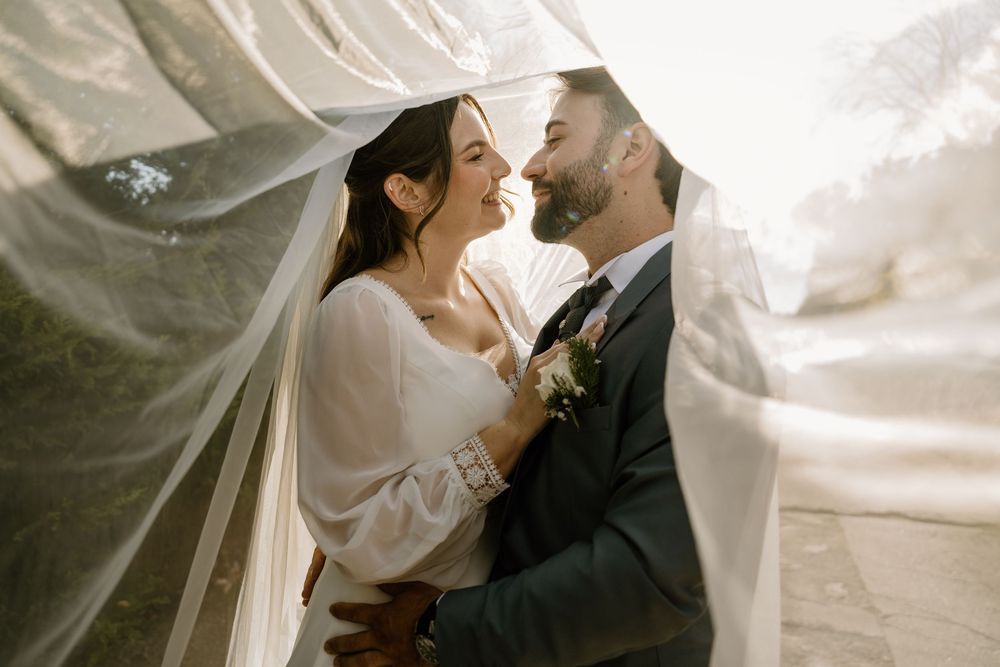 Smiling bride and groom embrace closely beneath a sheer flowing veil during golden-hour wedding portraits outdoors.