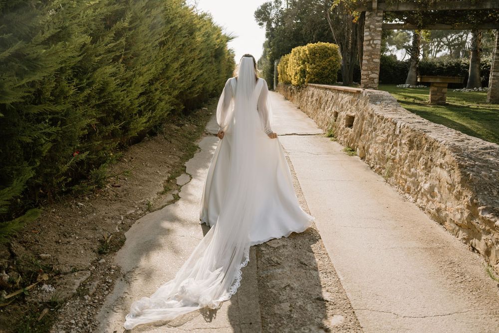 Bride in a long white gown and flowing veil walks away along a sunlit stone pathway lined with green hedges and an old stone wall.
