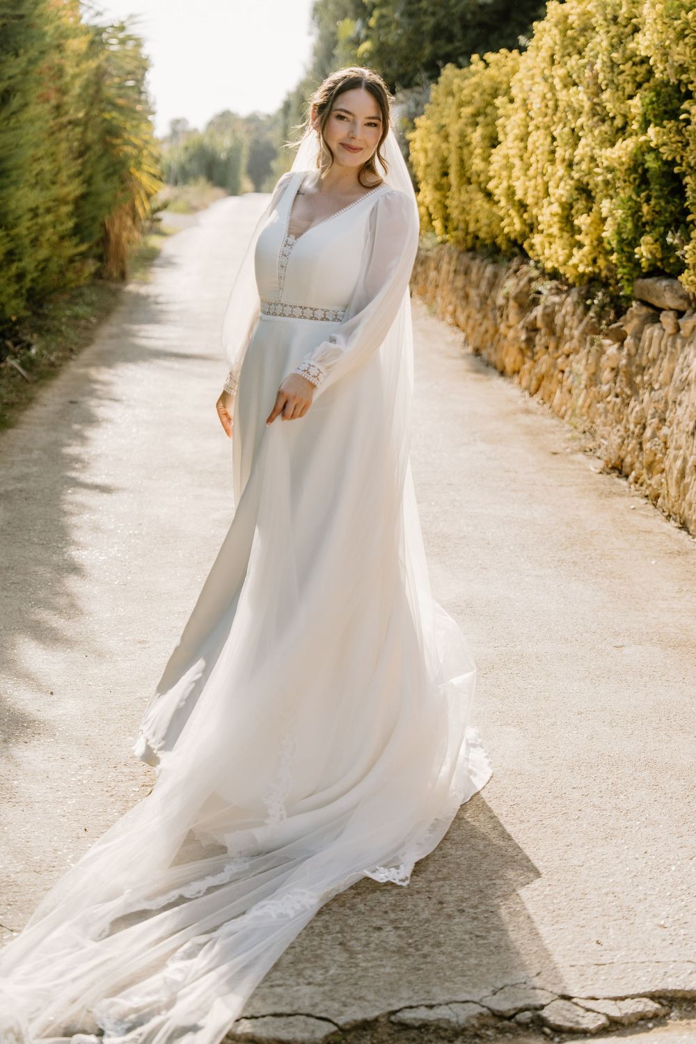 Smiling bride in an elegant long-sleeved white wedding dress with a long veil walks down a sunlit path lined with hedges.