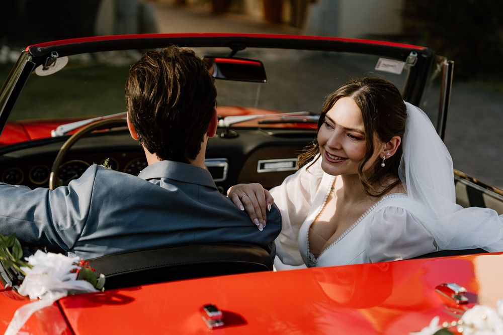 Smiling bride in a white dress and veil rests her hand on the groom's shoulder as they sit in a red convertible.