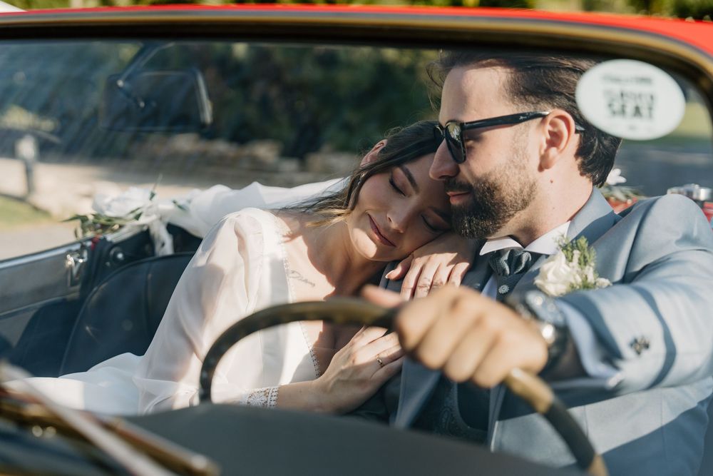 Bride in a white dress rests her head on the shoulder of a groom in sunglasses and gray suit as they sit in a vintage convertible.