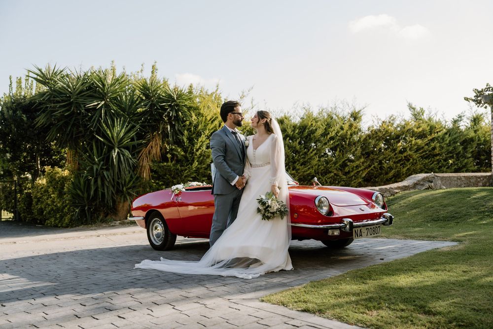 Bride and groom embrace on a paved path beside a vintage red convertible with a bouquet, surrounded by lush green plants.