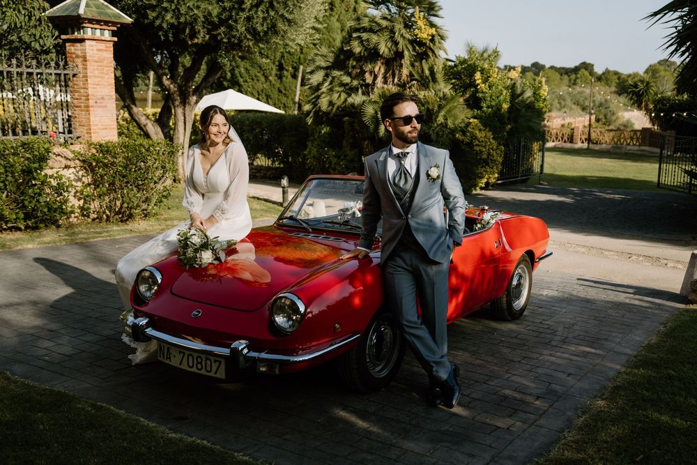 Bride in a white gown and groom in a gray suit pose beside a vintage red convertible on a sunny garden path in a wedding photo.