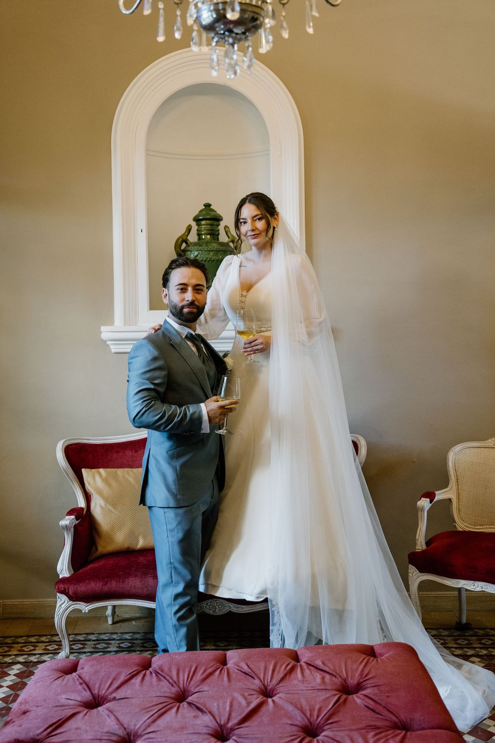 Bride in a white gown with a long veil and groom in a gray suit hold champagne glasses in an elegant room with red velvet chairs.