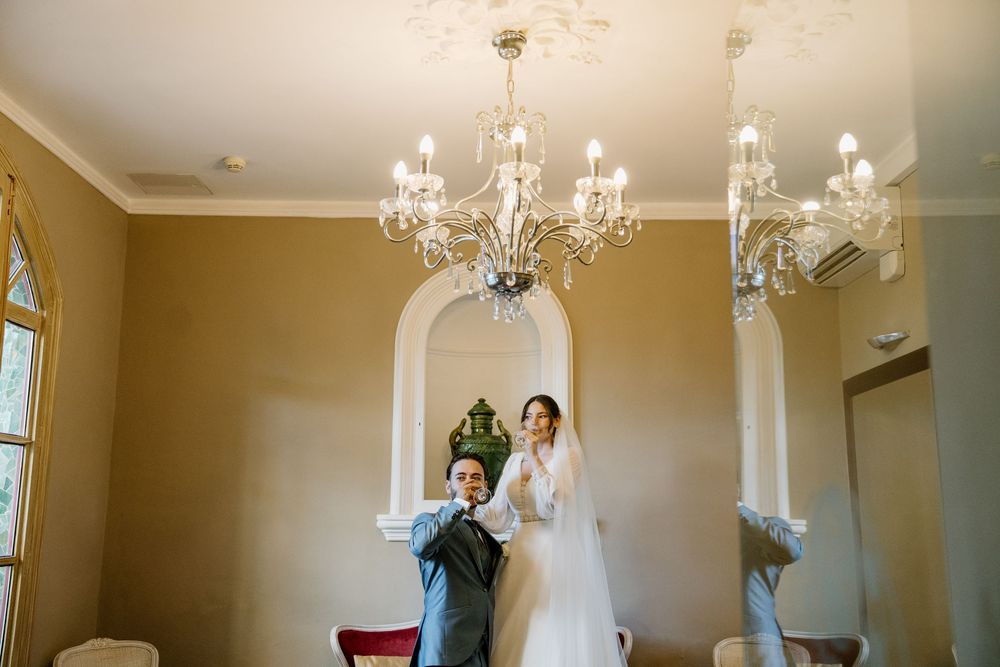 Bride and groom embrace in an elegant ballroom with crystal chandeliers, the bride in a veil and the groom in a gray suit.