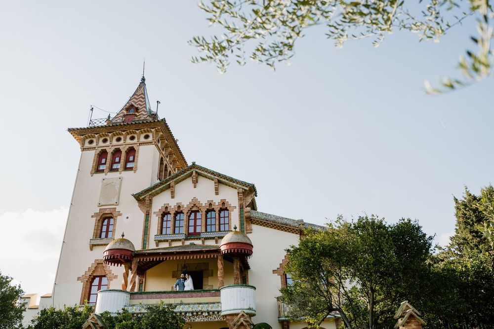 Ornate historic villa with a tall corner tower, arched windows and decorative balconies, surrounded by lush green trees under a soft sky.