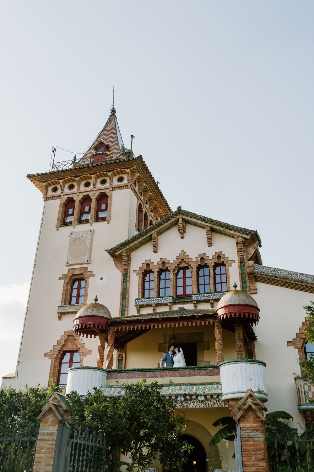 Couple embracing on the balcony of an ornate Catalan modernista villa with a tall tower, decorative brickwork, and red-trimmed turrets.